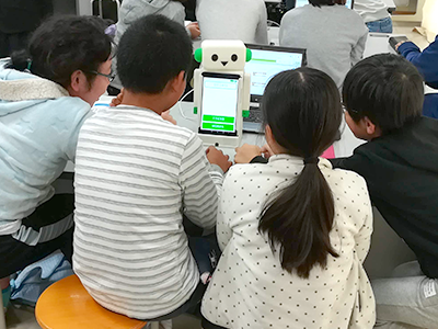 ロボットこくりのおなか部分のタブレットを操作している小学生のグループ